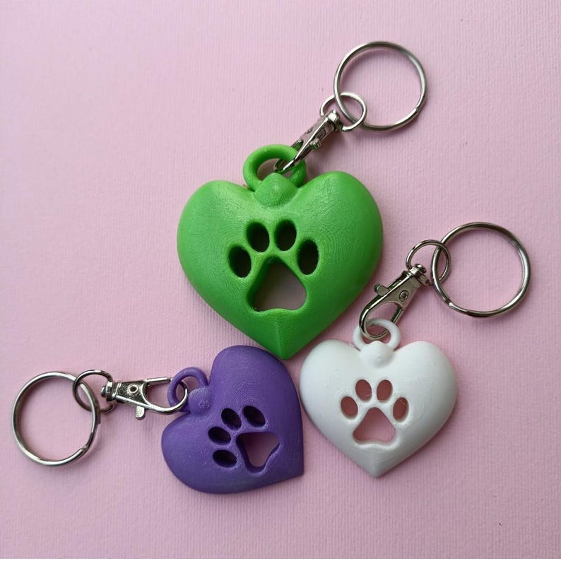 Puede incluir: Tres llaveros en forma de coraz&oacute;n en colores verde, morado y blanco. Cada llavero presenta un dise&ntilde;o de huella recortada en el centro y est&aacute; unido a una anilla de metal plateado con un cierre de mosquet&oacute;n para sujetar a las llaves.