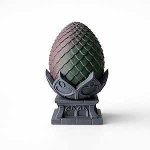 May include: A decorative dragon egg sculpture in shades of green and maroon, resting on a gray pedestal. The egg features a scale-like pattern. The pedestal has an ornate design, adding to the fantasy theme.