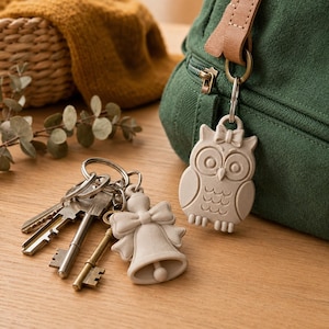 May include: A light beige owl-shaped keychain attached to a green fabric bag, alongside a separate metal keyring featuring a light beige bell-shaped charm and several silver and brass-colored metal keys resting on a wooden surface.