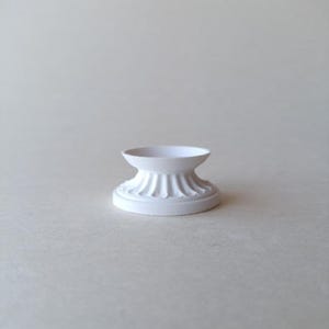 May include: A small, white, decorative pedestal with a fluted design. The pedestal has a round base and a shallow, bowl-shaped top. The object is made of a matte material and is suitable for displaying small items.