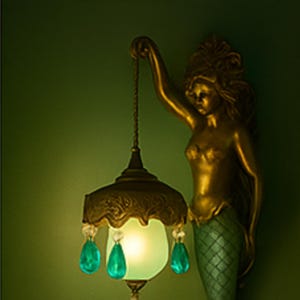 Op de afbeelding: Een gouden zeemeermin standbeeld houdt een hanglamp vast met een groene glazen kap en teal traanvormige kristallen. De lamp heeft een sierlijk metalen frame in antieke stijl. De zeemeermin heeft een groene geschubde staart.