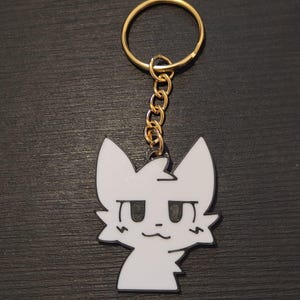 Può includere: Un portachiavi a forma di gatto bianco con contorno nero e anello dorato. Il gatto ha un design semplice e a cartoni animati con grandi occhi e una bocca piccola. Il portachiavi è realizzato in materiale lucido.