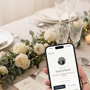 Könnte beinhalten: Ein Smartphone zeigt eine Hochzeitsgalerie-App mit den Namen Natali & Marko. Eine Menükarte mit QR-Code liegt auf einem Tisch mit weißen Rosen und Grün. Auf dem Telefon steht "Digital Wedding Camera - Your Live Gallery via QR Code."