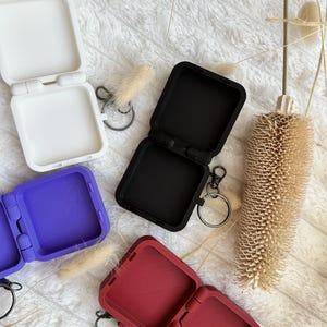 Puede incluir: Cuatro estuches cuadrados de silicona con tapas abatibles en blanco, negro, morado y rojo oscuro. Cada estuche presenta una base interior texturizada y un llavero de metal unido al lateral, diseñado para guardar objetos pequeños de forma segura.