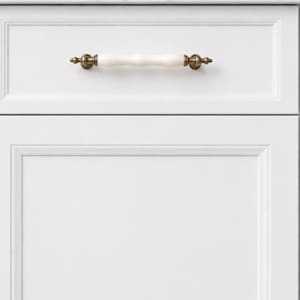 Puede incluir: Puerta de armario blanca con un panel decorativo y un cajón. El cajón tiene un tirador blanco y dorado. El armario es de color blanco brillante y los herrajes tienen un acabado de latón antiguo.
