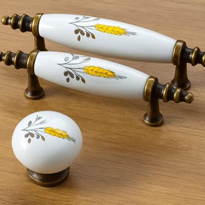 May include: White ceramic cabinet hardware with a wheat design. The set includes two pull handles and a round knob, all with bronze-colored metal accents. The wheat design is in yellow and brown. The hardware is displayed on a wooden surface.