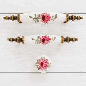 May include: A set of three white ceramic cabinet hardware pieces. Two are elongated handles, and one is a round knob. Each features a hand-painted pink flower design with green leaves and gold-colored metal accents.