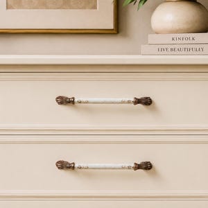 Peut inclure: Commode crème avec deux tiroirs, chacun avec une poignée décorative. Les poignées ont un centre blanc semblable à du marbre et des extrémités ornées de couleur bronze. La commode est placée contre un mur aux tons neutres.