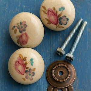 Può includere: Quattro pomelli per mobili in ceramica color crema con disegni floreali rosa, blu e oro. Sono inclusi anche due bulloni argentati e due rondelle in bronzo. I pomelli hanno una finitura craquelé, su sfondo blu.
