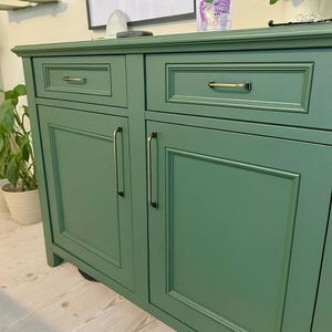 May include: A green cabinet with two drawers and two doors. The cabinet has gold-colored handles and a framed design on the doors and drawers. The cabinet is painted in a solid green color and is placed against a wall.