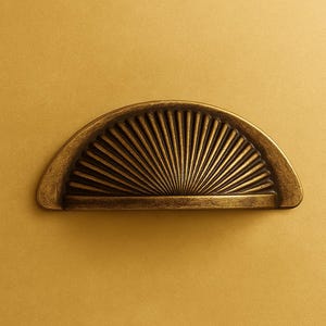 May include: A bronze-coloured, half-moon cabinet pull with a fan design. The hardware is set against a gold background. The pull is likely for a drawer or cabinet door, and is made of metal.