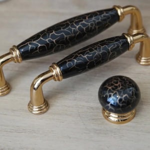 Pode incluir: Conjunto de duas puxadores de armário pretos e dourados e um puxador a condizer. As puxadores têm um centro preto com padrão craquelado e ferragens douradas. O puxador é uma esfera com o mesmo padrão craquelado e base dourada.