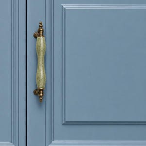 May include: A vertical cabinet handle featuring a central pale green ceramic grip with a subtle crackle glaze finish, secured by antique brass-toned metal mounts, attached to a flat-panel cabinet door painted in a solid matte dusty blue color.