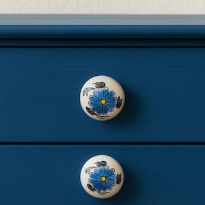 Puede incluir: Primer plano de una cómoda azul oscuro con dos cajones. Cada cajón tiene un pomo redondo blanco con un diseño floral azul. Los pomos tienen un centro amarillo y detalles en negro. La cómoda está pintada de un color azul oscuro sólido.
