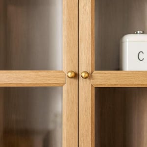 May include: Close-up of a wooden cabinet with glass doors. The cabinet has a light oak finish and brass-coloured knobs. A white container with the letter "C" is visible inside the cabinet.