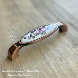 Peut inclure: Tirette de tiroir florale peinte à la main avec une finition cuivrée. Le centre en céramique blanche présente un motif floral dans les tons de violet, de rouge et de vert. Le texte "Hand-Painted Floral Drawer Pull" est visible.