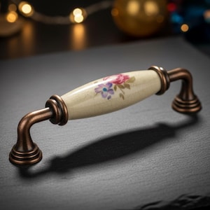 May include: A cabinet handle featuring a cream ceramic centre with a floral design in pink and purple. The curved supports and ends are dark bronze, offering a vintage appearance. The handle is suitable for cabinets.