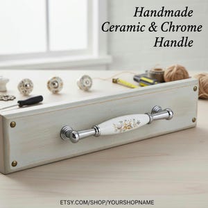 Pode incluir: Uma puxador de cerâmica e cromo feito à mão com um centro de cerâmica branca com um design floral dourado. O puxador é montado numa superfície de madeira cinza clara. Vários puxadores decorativos e ferramentas também são visíveis.