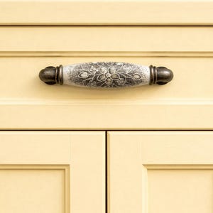 May include: Close-up of a cream-colored cabinet with a decorative handle. The handle is oval-shaped with a floral design in shades of gray and white. The ends of the handle are dark bronze.