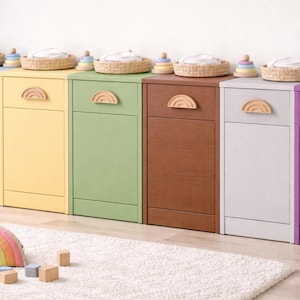 Puede incluir: Seis armarios de almacenamiento coloridos en azul, amarillo, verde, marrón, blanco y morado. Cada armario tiene un tirador de madera en forma de arcoíris. Encima hay cestas tejidas y juguetes de madera apilables. Una almohada en forma de arcoíris y bloques de madera en el suelo.