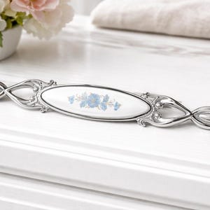 May include: A decorative cabinet pull with a white oval centre featuring a blue floral design. The pull is made of silver-toned metal with ornate detailing and is approximately 15 cm long.