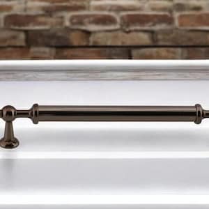 May include: A dark bronze cabinet pull with decorative end details. The hardware is a long, straight bar with a cylindrical shape and is mounted on a white surface. The background features a brick wall.