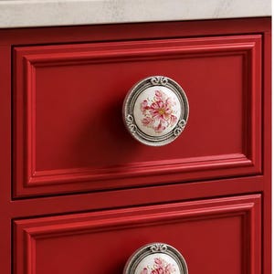 May include: A close-up of a red cabinet with two drawers, each featuring a decorative floral knob. The knobs have a white background with pink and red flower designs, set in a silver-coloured frame.
