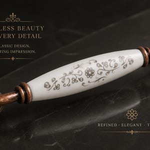 May include: A decorative cabinet pull with a white ceramic center and bronze-colored metal ends. The white section features a floral pattern. The image includes the text "Timeless beauty in every detail" and "Refined, Elegant, Timeless."