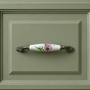 May include: A decorative cabinet pull with a white ceramic handle featuring a floral design in pink and green. The ends are bronze-colored with a detailed, ornate design. The pull is mounted on a sage green cabinet door.