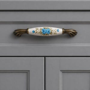 May include: Close-up of a gray cabinet door with a decorative handle. The handle has a white center with blue floral designs and brass-colored ends. The cabinet door is a light gray color.