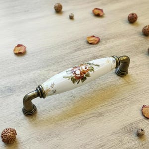 Peut inclure: Tirette de meuble ovale avec un centre en céramique blanche orné d'un motif floral dans les tons de marron, rouge et vert. Les extrémités et la quincaillerie sont de couleur bronze. Quincaillerie décorative pour meubles.