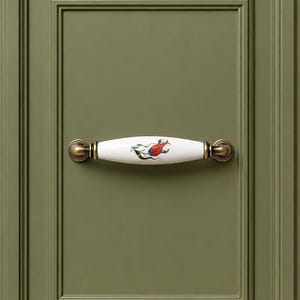 May include: A white ceramic cabinet pull with a floral design in red, green, and blue, mounted on a green cabinet door. The pull has brass-colored end caps and a curved shape, adding a decorative touch to the cabinetry.