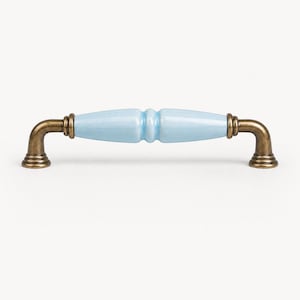 May include: A cabinet pull with a light blue ceramic center and antique brass-colored metal hardware. The handle has a cylindrical shape with a slight hourglass design in the center. The hardware has a curved, right-angle shape.