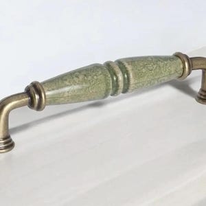 May include: A cabinet handle with a green and cream ceramic centre and antique brass finish. The handle has a decorative, textured design and is mounted on a white surface. Suitable for drawers and cabinets.