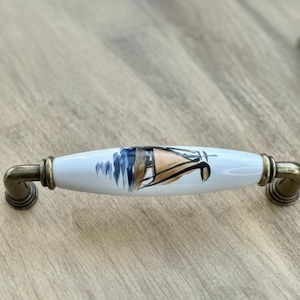 May include: Three white ceramic cabinet pulls with a sailboat design in blue and brown. The pulls have a brass finish and are arranged on a light wood surface. Decorative hardware for furniture.