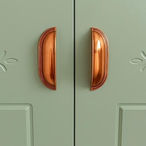 Może przedstawiać: Zbliżenie pary drzwi szafki pomalowanych na miękki zielony odcień. Każde drzwi mają miedziany uchwyt w kształcie półksiężyca. Uchwyty mają żebrowany wzór i są zamontowane na drzwiach. Widoczne są dekoracyjne wycięcia w kształcie kwiatów.