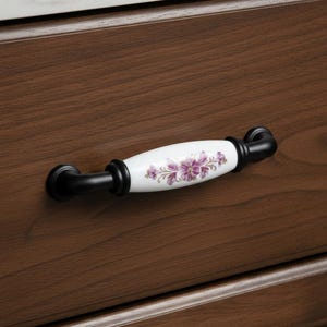 Peut inclure: Poignée de meuble avec un centre en céramique blanche orné d'un motif floral violet. La poignée est dotée d'une quincaillerie en métal noir et est montée sur une porte d'armoire en bois brun foncé. La poignée est un élément décoratif.
