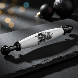 Op de afbeelding: Een witte kastgreep met een zwart bloemmotief. De greep heeft een cilindrisch midden met een zwart bloemmotief en zwarte uiteinden. De hardware staat tegen een donkere achtergrond met vage lichten.