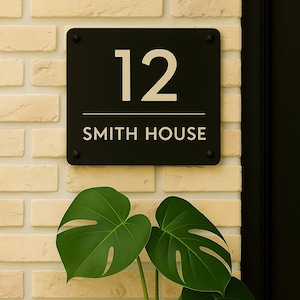 May include: A black rectangular house sign displaying the number "12" and the words "SMITH HOUSE" in white. The sign is fixed to a light beige brick wall. A green Monstera plant with large leaves is in the foreground.