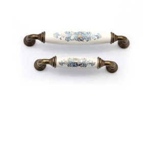 May include: Two cabinet handles. The handles have a white ceramic centre with a blue floral design. The ends are a bronze colour. One handle is smaller than the other.