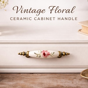 Op de afbeelding: Witte keramische kastgreep met een bloemmotief. De greep heeft een crèmekleurig midden met een roze bloem en groene bladeren. De uiteinden zijn goudkleurig. De tekst "Vintage Floral CERAMIC CABINET HANDLE" staat erboven.