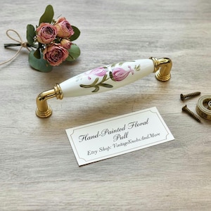 May include: A hand-painted floral pull with a white ceramic handle and gold-toned metal accents. The handle features pink floral designs. A business card reads "Hand-Painted Floral Pull".