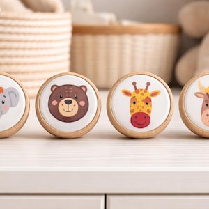 Peut inclure: Quatre boutons de meuble en bois avec des visages d'animaux : un éléphant, un ours, une girafe et une vache. Chaque bouton a un fond blanc avec un visage d'animal de dessin animé coloré et un bord en bois naturel. Les boutons sont ronds et d'environ 3,8 cm de diamètre.