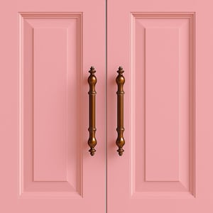 Könnte beinhalten: Nahaufnahme von zwei rosa Schranktüren mit verzierten, bronzefarbenen Griffen. Die Türen haben ein eingelassenes Paneeldesign und eine glatte, matte Oberfläche, was eine klassische und elegante Ästhetik schafft.
