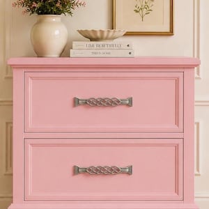 May include: A two-drawer pink dresser with silver-toned hardware. The dresser is topped with a white vase of pink flowers, a decorative bowl, and two books. A framed floral print hangs on the wall above.