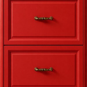 Pode incluir: Um armário vermelho de duas gavetas com puxadores cor de latão. O armário tem um design clássico com painéis embutidos em cada gaveta. As puxadores são horizontais e têm um design decorativo e ornamentado.