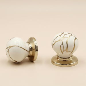 以下が含まれることがあります： 白と金のキャビネットノブが2つ。ノブは、金色の静脈を持つ丸い大理石のようなデザインです。ベースとハードウェアは金色です。ノブは、キャビネットまたは引き出し用である可能性があります。