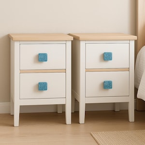 Op de afbeelding: Twee witte nachtkastjes met elk twee lades, met licht houten bladen en blauwe vierkante ladeknopen. De nachtkastjes staan naast elkaar. De nachtkastjes zijn een slaapkamermeubel. Hoogte ongeveer 60 cm.