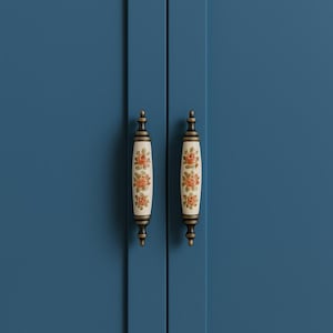 May include: Two cabinet handles with a cream-colored, floral design. The handles are cylindrical with brass-colored hardware and are set against a blue cabinet door.