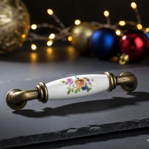 May include: A cabinet pull with a white ceramic center featuring a floral design in pink, yellow, and blue. The ends are antique brass. The hardware is designed for furniture and home decor.